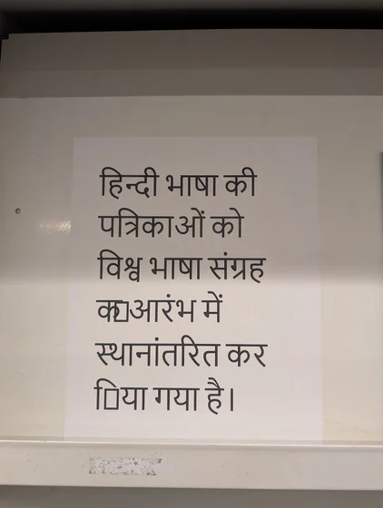 A photograph of a white sheet of paper placed on a light-colored shelf. On the paper is printed Hindi text that reads:
हिन्दी भाषा की
पत्रिकाओं को
विश्व भाषा संग्रह
क▯ओरंभ में
स्थानांतरित कर
▯िया गया है।

Two vertical rectangles (tofu symbols) are found in the text.