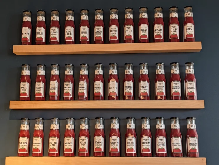 A wall of 36 ketchup-looking bottles, numbered from 01 to 36. Each is labelled with “FOOD HALL 6 2023” and a different main text:
01: ALT + F4
02: PRESS ANY KEY TO CONTINUE
03: 175,000+ Employees
04: R:170 G:37 B:0
05: 18 BILLION CORTANA QUESTIONS
06: LUNCH REBOOT
07: AI FOR EARTH
08: CTL + Y
09: NOV. 28 2017
10: UPDATE 87% COMPLETE
11: CTL + ALT + TAB
12: OPEN SAUCE CODE
13: MAR. 13 1996
14: 100% CARBON NEUTRAL GLOBALLY
15: NEW TECHNOLOGY PARADIGM
16: SOMETHING HAPPENED
17: CORTANA, TELL ME A …