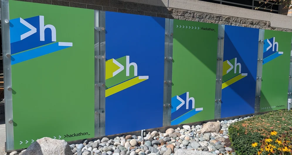 A wall of 5 posters in a green-blue-yellow palette, each has “>h_” written in large size.
