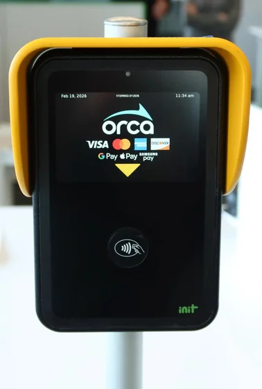 Close-up of a black Orca contactless payment terminal in a bright yellow frame. The screen shows the Orca logo with Visa, Mastercard, Discover, Google Pay, Apple Pay, and Samsung Pay logos, plus a tap-to-pay icon below.