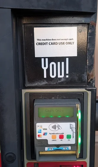 Payment modules of a vending machine. 
The top module is replaced by a sticker that reads “This machine does not accept cash. CREDIT CARD USE ONLY.”
The bottom module is a card reader with a screen that shows “Cash Only.”

Between these two is a sticker that reads “You!”