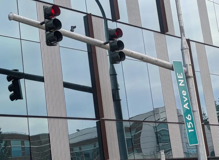 A street sign reading "156 Ave NE" is mounted on a metal pole next to two traffic lights. Only the right side of the sign is mounted, and the sign is dangling on the mount point.