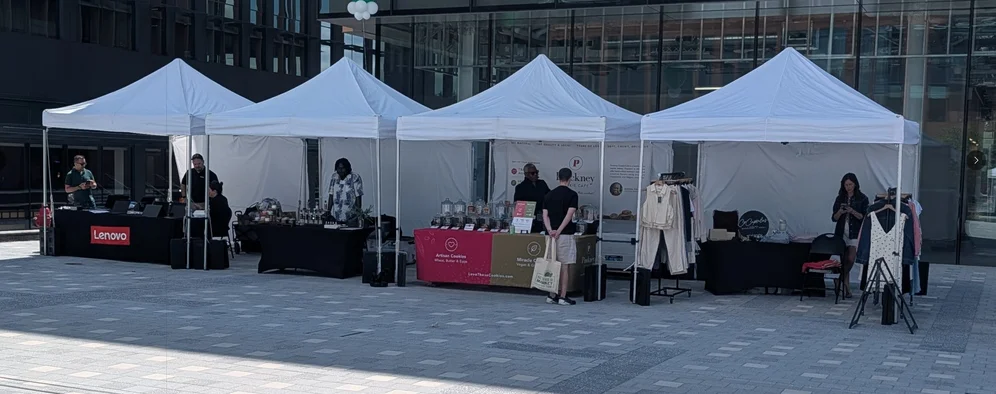 Photo of 4 temporary tent booths in a square. Each is selling, from right to left, clothing, cookies, essential oil, Lenovo laptops.