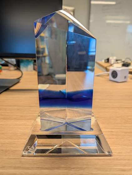 A faceted clear crystal trophy with a deep blue gradient at its center, resting on a wide square glass base. The award is shaped like a tall pillar with geometric, angled cuts at the top that catch the light. It sits on a light wooden desk in an office setting.